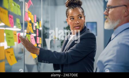 Senior Manager Talking to Experienced Businessman while Creating Time Management Plan on Office Wall. Colleagues Discuss Commercial, Financial and Marketing Projects. Specialists Work in Diverse Team. Stock Photo