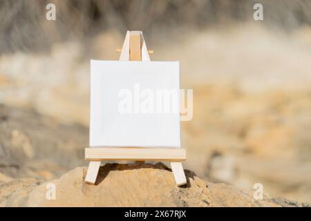 Mockup. White mini canvas on small wooden easel stand standing outdoors with blurred nature background Stock Photo