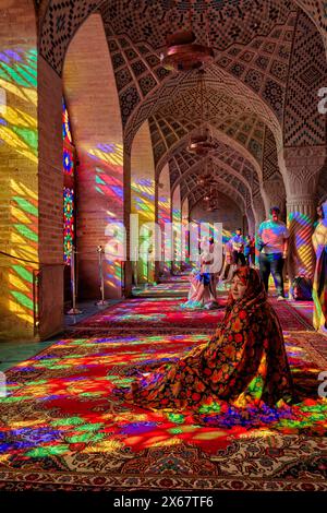 A woman sits in the light from multi-colored stained-glass windows inside the 19th century Nasir al-Mulk Mosque, aka the Pink Mosque. Shiraz, Iran. Stock Photo