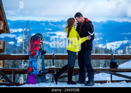 Young caucasian straight couple in love hugging each other almost kissing on a snowy ski resort near colorful snowboards standing aside, engagement se Stock Photo
