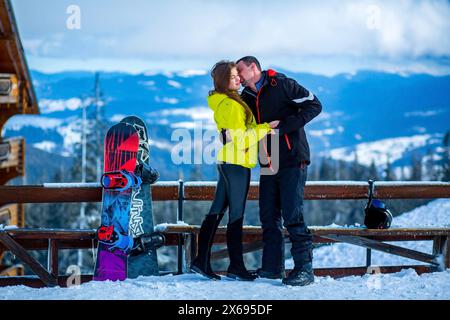 Young caucasian straight couple in love hugging each other almost kissing on a snowy ski resort near colorful snowboards standing aside, engagement se Stock Photo