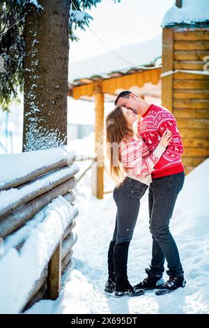 Engagement session, love story of a young straight couple hugging and almost kissing in Christmas red sweaters and jeans in snowy landscape near a woo Stock Photo