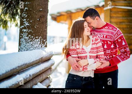 Engagement session, love story of a young straight couple hugging and almost kissing in Christmas red sweaters and jeans in snowy landscape near a woo Stock Photo