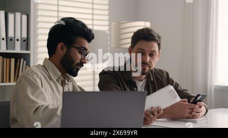 Multiethnic employees talk to client two diverse men businessmen discuss project on laptop business coach mentor teaching intern using mobile phone Stock Photo