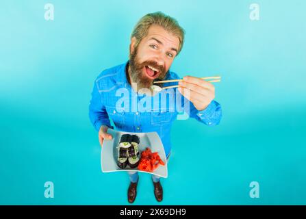 Bearded man eating tasty sushi roll with bamboo chopsticks. Handsome male in denim shirt with plate of sushi rolls and pickled ginger. Sushi delivery Stock Photo