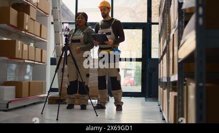 Warehouse head of operations and coworker using smartphone placed on tripod to make training video for interns. Directors film themselves in fulfillment center showing trainees how to store boxes Stock Photo