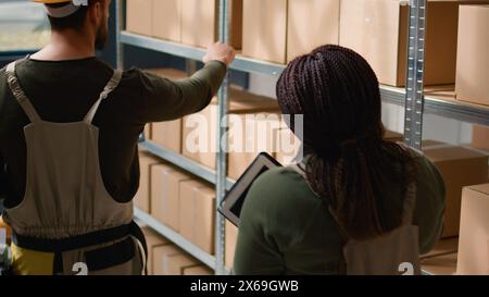 Skillful staff members preparing warehouse orders for delivery, using laptop and tablet to verify shipping informations, crosschecking online data with stock labels in storage establishment Stock Photo
