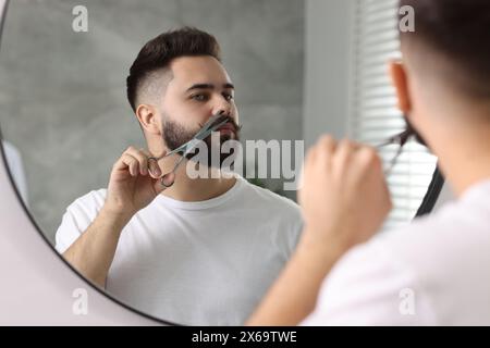 Handsome young man trimming mustache with scissors near mirror in bathroom Stock Photo