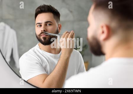 Handsome young man trimming mustache with scissors near mirror in bathroom Stock Photo