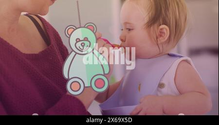 Image of teddy bear icon over caucasian mother feeding her baby with spoon Stock Photo