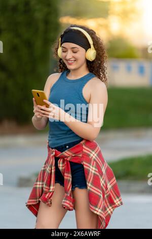 Joyful curly-haired woman in blue tank top using a smartphone with headphones outdoors in a park at sunset, with a plaid shirt tied around her waist.  Stock Photo