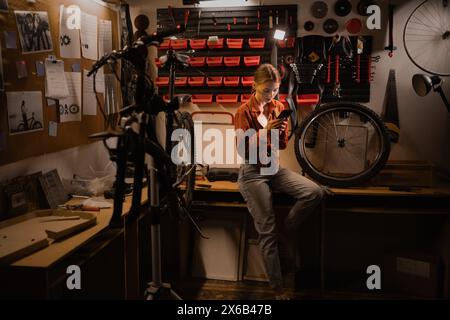 Young woman looking on phone searching help on repairing damaged bike in workshop or garage. Lady on telephone call with bicycle engineer Stock Photo
