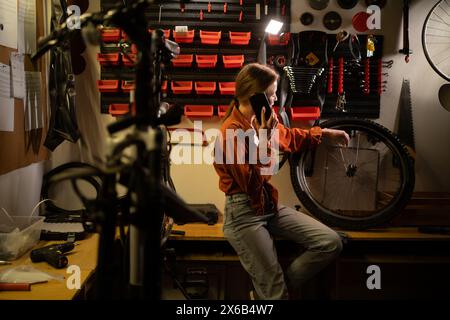 Bicycle maintenance and repair concept. Young blonde woman working in garage calling on mobile phone consulting with mechanic Stock Photo