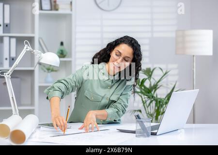 A professional Hispanic woman in a modern office setting meticulously works on architectural drawings beside her laptop, displaying focus and expertise. Stock Photo