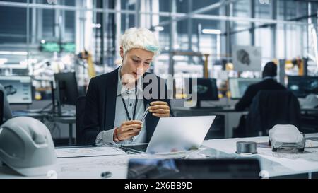 Young Beautiful Engineer Working and Managing Projects on Laptop Computer in an Office at Car Assembly Plant. Industrial Specialist Working on Vehicle Parts in Technological Development Facility. Stock Photo
