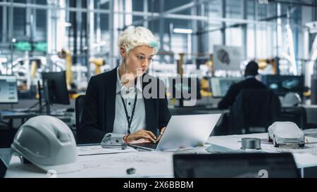 Young Beautiful Engineer Working and Managing Projects on Laptop Computer in an Office at Car Assembly Plant. Industrial Specialist Working on Vehicle Parts in Technological Development Facility. Stock Photo