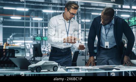 Two Automotive Engineers Working in Office at Car Factory. Industrial Designer Shows Conceptual Electric Engine Parts to Colleague, Discussing Different Applications in Technological Laboratory. Stock Photo