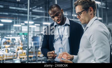 Two Diverse Automotive Industrial Engineers Talking About Vehicle Production while Standing in Office at a Car Assembly Plant. Industrial Specialists Discuss Work Projects on Tablet Computer. Stock Photo