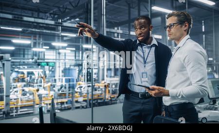Two Diverse Automotive Industrial Engineers Talking About Vehicle Production while Standing in Office at a Car Assembly Plant. Industrial Specialists Discuss Work Projects on Tablet Computer. Stock Photo