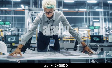 Young Female Engineer Looking at a Technical Blueprint at Work in an Office at Car Assembly Plant. Industrial Specialist Working on Vehicle Parts in Technological Development Facility. Stock Photo
