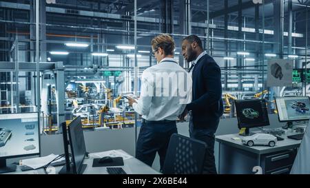 Two Diverse Automotive Industrial Engineers Talking About Vehicle Production while Standing in Office at a Car Assembly Plant. Industrial Specialists Discuss Work Projects on Tablet Computer. Stock Photo