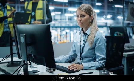 Car Factory Office: Portrait of a Concentrated Female Engineer Working on Desktop Computer. Team of Technicians in Automated Robot Arm Assembly Line Manufacturing Electronics Stock Photo