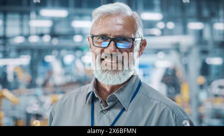 Car Factory Office: Portrait of Senior White Male Chief Engineer Looking at Camera and Smiling. Professional Technician in Automated Robot Arm Assembly Line Manufacturing in High-Tech Facility Stock Photo