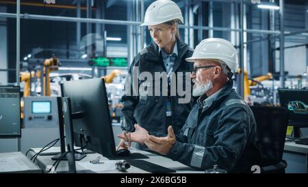 Car Factory Office: Female Manager Talks to Male Automotive Engineer Working on Computer. Automated Robot Arm Assembly Line Manufacturing Electric Vehicles. Technicians Monitoring Conveyor Production Stock Photo