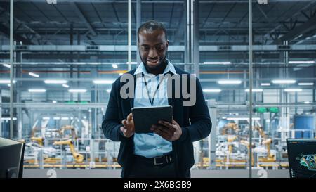 Car Factory Office: Portrait of Successful Black Male Chief Engineer Using Tablet Computer in Automated Robot Arm Assembly Line Manufacturing High-Tech Electric Vehicles. Medium Shot Stock Photo