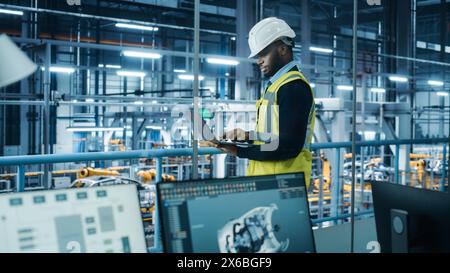 Car Factory: Professional Male Automotive Engineer Wearing Hard Hat, Walking, Using Laptop. Monitoring, Control, Equipment Production. Automated Robot Arm Assembly Line Manufacturing Electric Vehicles Stock Photo