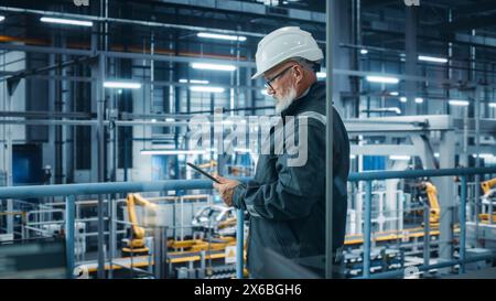 Car Factory: Male Automotive Engineer Wearing Hard Hat, Standing Using Tablet Computer. Monitoring, Control, Equipment Production. Automated Robot Arm Assembly Line Manufacturing Electric Vehicles Stock Photo
