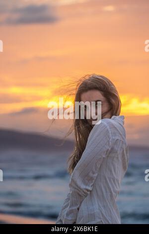 Woman gazes softly over her shoulder, the sunset illuminating her hair and features with warm hues, as the tranquil ocean serves as an idyllic backdro Stock Photo