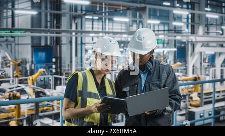 Male Specialist and Female Car Factory Engineer Talking and Working on Laptop and Tablet Computers. Automotive Industry 4.0 Facility Working on Vehicle Production. Diversity on Assembly Plant. Stock Photo