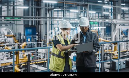 Male Specialist and Female Car Factory Engineer Talking and Working on Laptop and Tablet Computers. Automotive Industry 4.0 Facility Working on Vehicle Production. Diversity on Assembly Plant. Stock Photo