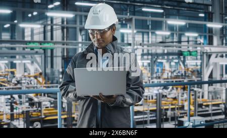 Portrait of Automotive Industry Engineer in Safety Uniform Using Laptop at Car Factory Facility. Happy Assembly Plant Multiethnic Man Specialist Working on Manufacturing Modern Electric Vehicles. Stock Photo