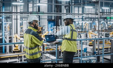 Two Car Factory Specialists in High Visibility Vests Using Laptop Computer. Engineers Discussing Automotive Industrial Manufacturing Facility Process of Vehicle Production. Automated Assembly Plant. Stock Photo