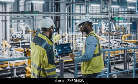 Two Car Factory Specialists in High Visibility Vests Using Laptop Computer. Engineers Discussing Automotive Industrial Manufacturing Facility Process of Vehicle Production. Automated Assembly Plant. Stock Photo