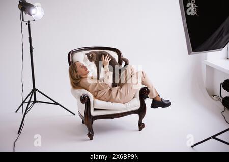 Female blogger, blonde on chair with domestic cat scottish straight in white photo studio. Dressed in a formal beige trouser suit Stock Photo