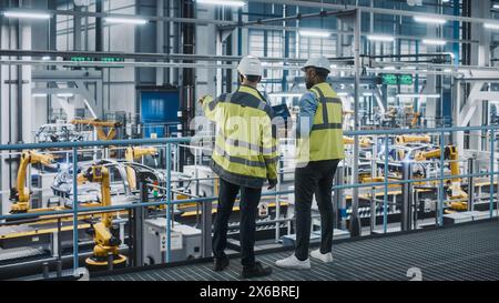 Two Car Factory Specialists in High Visibility Vests Using Laptop Computer. Engineers Discussing Automotive Industrial Manufacturing Facility Process of Vehicle Production. Automated Assembly Plant. Stock Photo