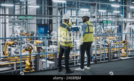 Two Car Factory Specialists in High Visibility Vests Using Laptop Computer. Engineers Discussing Automotive Industrial Manufacturing Facility Process of Vehicle Production. Automated Assembly Plant. Stock Photo