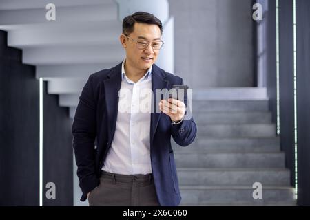 Asian mature businessman using his smartphone while standing in a modern office building with a confident expression. Stock Photo