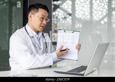 Asian mature male doctor explaining a medical report during a video consultation using a laptop in a modern office. Stock Photo
