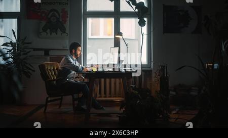 Young Handsome Man Works on a Laptop Computer in Creative Agency in Loft Office. He Writes Down a Note with Pen. Renovated Stylish Decor with House Plants, Artistic Posters and Big Rounded Windows. Stock Photo