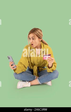 Full body view caucasian blonde shocked young woman sit floor holding mobile phone and credit card looking smartphone screen. Online shopping concept Stock Photo