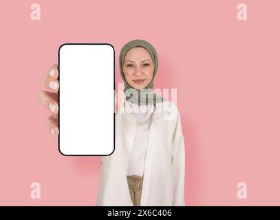 portrait of Muslim woman showing empty blank white screen mobile phone mock up. Application advertisement concept idea image. Isolated pink background Stock Photo