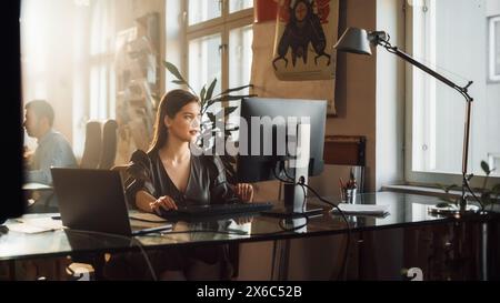 Two Stylish Employees Working on Computers in Creative Agency in Loft Office. Beautiful Manager Typing Correspondence Emails. Sunny Renovated Space with Plants, Artistic Posters and Big Windows. Stock Photo