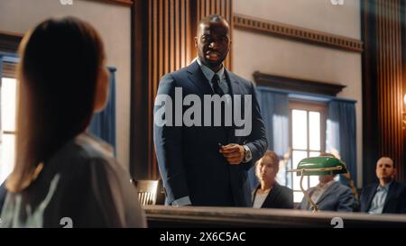 Court of Justice and Law Trial: Male Public Defender Presenting Case, Asking Female Witness in Front of Judge and Jury. African American Attorney Lawyer Protecting Client Against Crime, Injustice. Stock Photo