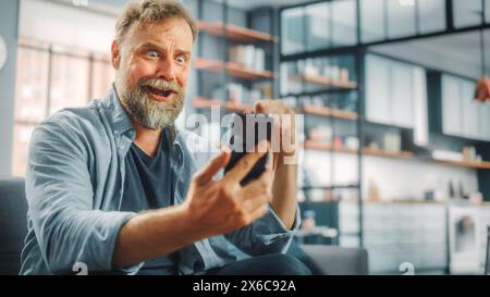 Charismatic Bearded Man Sitting on a Couch Watches Game on TV, uses Smartphone App for Score, Online Bet, Statistics, Celebrates Victory when Team Wins Championship. Happy Fan Watches Sport Stock Photo