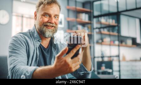Charismatic Bearded Man Sitting on a Couch Watches Game on TV, uses Smartphone App for Score, Online Bet, Statistics, Celebrates Victory when Team Wins Championship. Happy Fan Watches Sport Stock Photo