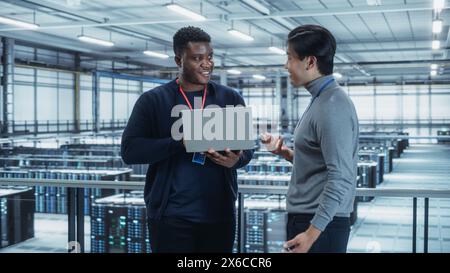 Two Multiethnic Male Data Center System Administrators and IT Specialists Talk, Use Laptop. Information Technology Engineers work on Cyber Security Network Protection in Cloud Computing Server Farm. Stock Photo
